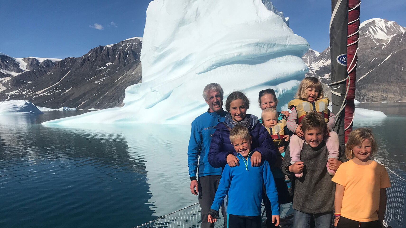 Familie Schw&ouml;rer posiert gemeinsam auf dem Deck ihres TOPtoTOP Expeditionssegelbootes vor einem m&auml;chtigen Eisberg in einem gr&ouml;nl&auml;ndischen Fjord &ndash; Eltern und six Kinder auf einer Klimaschutzreise