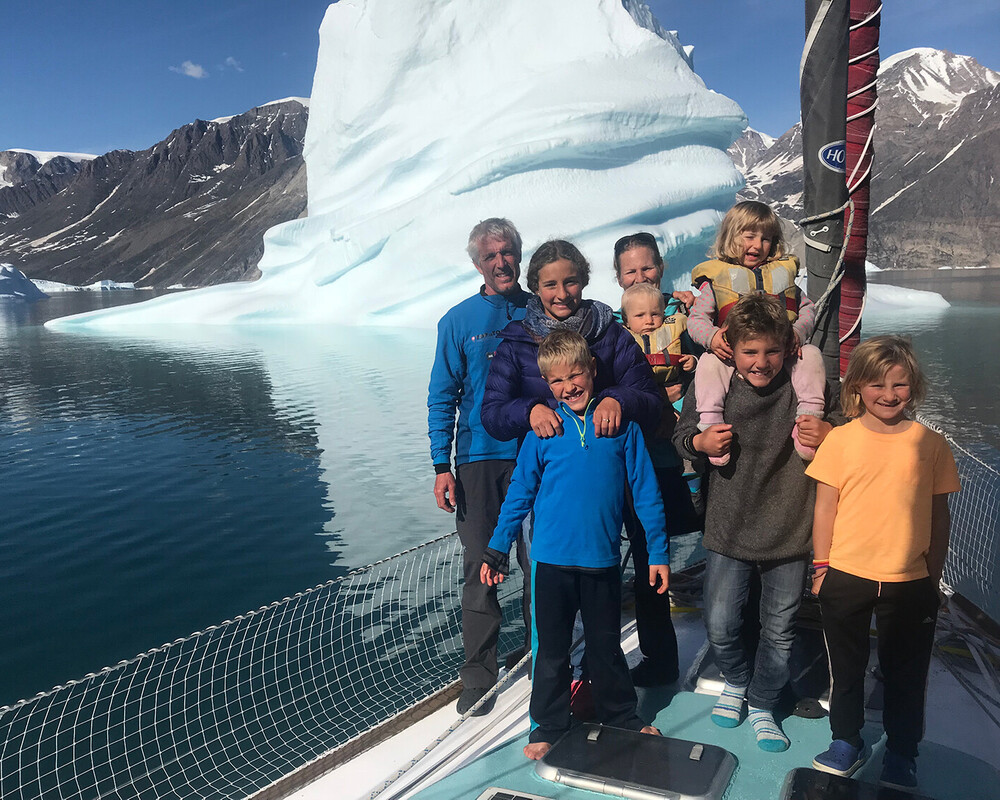 Familie Schw&ouml;rer posiert gemeinsam auf dem Deck ihres TOPtoTOP Expeditionssegelbootes vor einem m&auml;chtigen Eisberg in einem gr&ouml;nl&auml;ndischen Fjord &ndash; Eltern und six Kinder auf einer Klimaschutzreise
